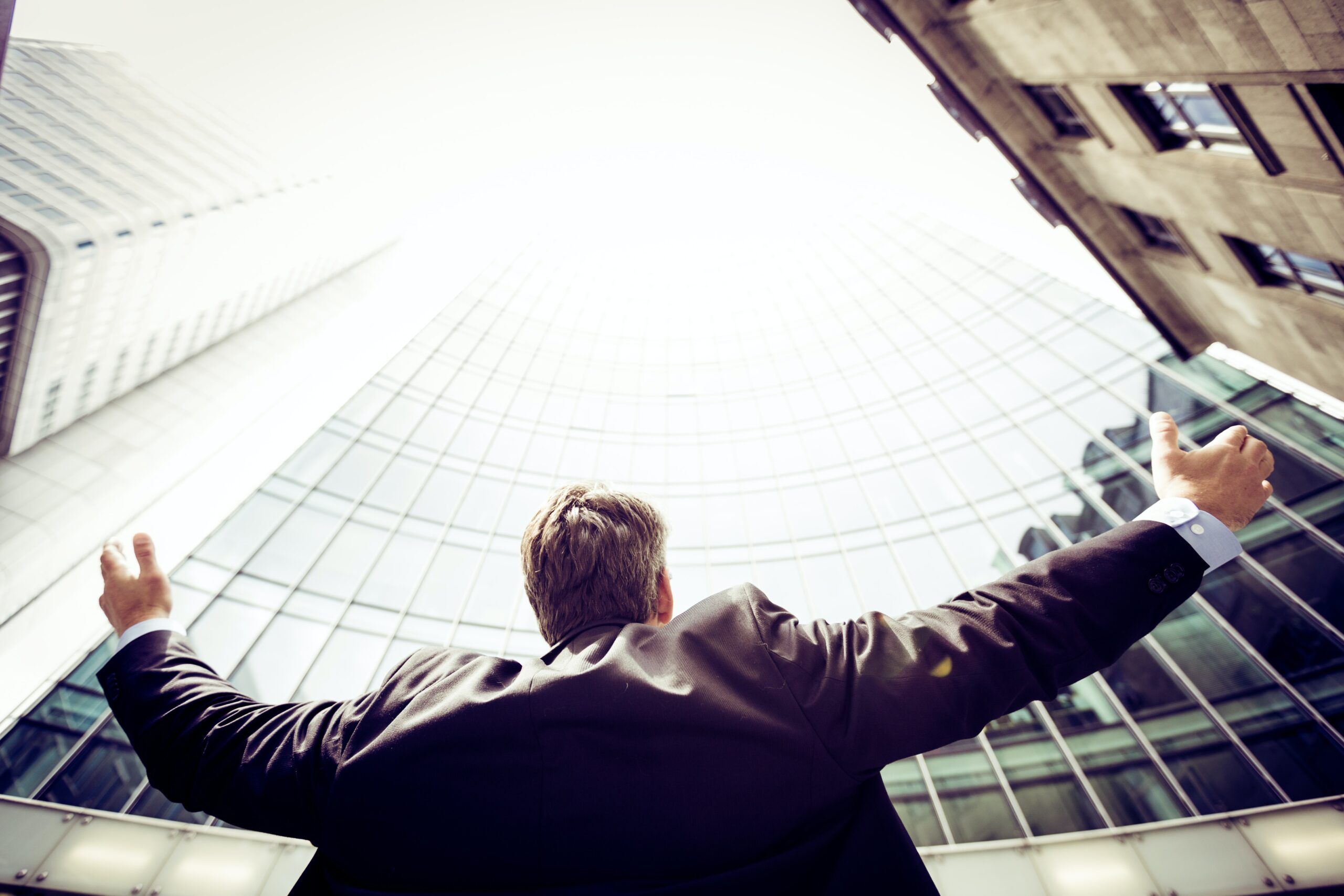 A business owner in a suit stands with his back to the camera, arms raised in a victorious or celebratory gesture, against the backdrop of modern high-rise buildings.