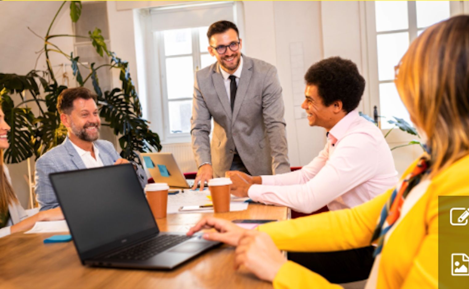 Four people sit at a table with laptops, papers, and coffee cups during a Leadership MasterCLASS meeting. One person stands and smiles while speaking to the group. Large plants and windows are visible in the background.