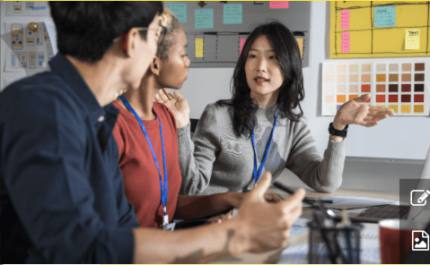 Three people sit at a desk in an office, engaged in discussion during a Marketing Training session. One woman gestures with her hand while talking. Color swatches and sticky notes are visible on the wall behind them.