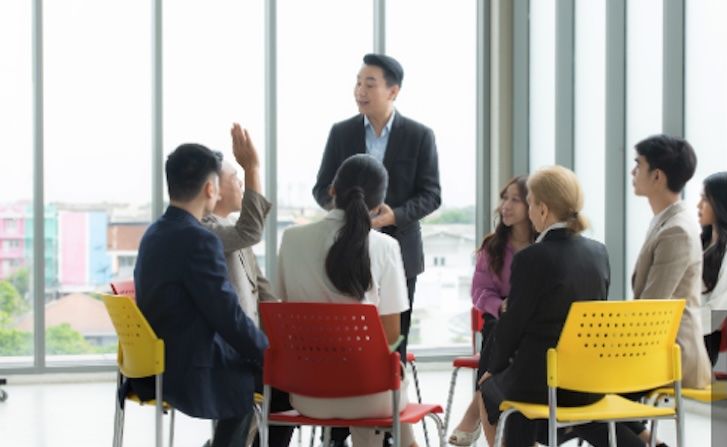 A man in a suit stands and speaks to a group of seven people seated in a circle of colorful chairs, with one person raising their hand—perhaps engaging in an interactive Sales MasterCLASS. Large windows are in the background.